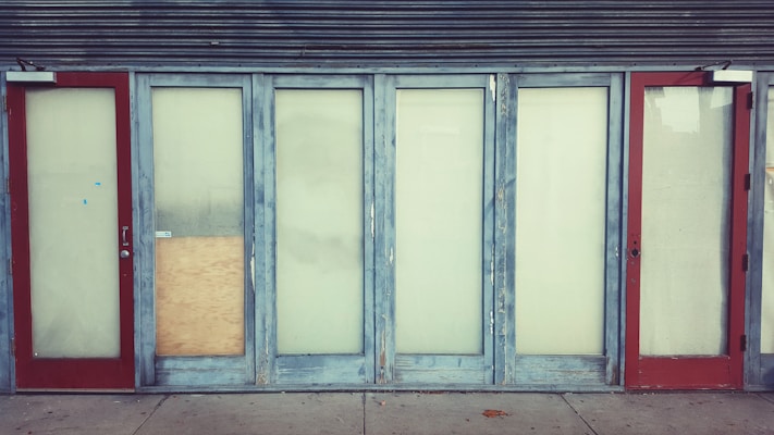 A row of closed doors with frosted glass panels set in a weathered blue frame. The doors are evenly spaced along the front of a building, with two red doors on either end. One of the central panels is covered with a piece of plywood.