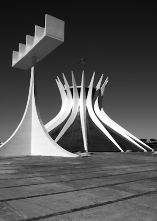 A striking architectural structure with a modern and abstract design. The building features tall, curved pillars reaching towards the sky, resembling a crown or a series of spikes. The foreground has a paved area that complements the futuristic and minimalist design.