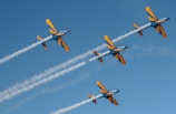 Four aerobatic planes fly in formation against a clear blue sky, leaving trails of smoke behind them. The planes are painted in yellow and blue colors.