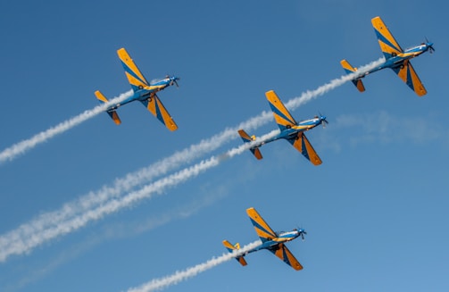 Four aerobatic planes fly in formation against a clear blue sky, leaving trails of smoke behind them. The planes are painted in yellow and blue colors.