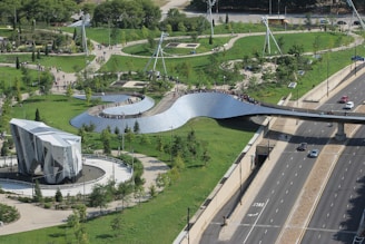 A large, modern park with a winding metallic pedestrian bridge spanning across a highway. The area is surrounded by lush green lawns, trees, and walking paths. There is a distinctive, geometric-shaped climbing structure situated in the park. People are seen walking and enjoying the outdoors.