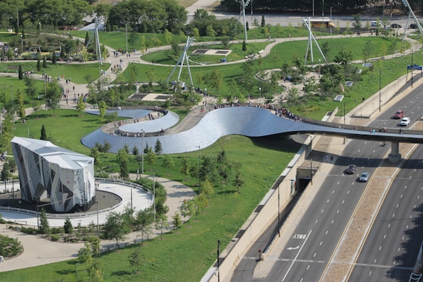 A large, modern park with a winding metallic pedestrian bridge spanning across a highway. The area is surrounded by lush green lawns, trees, and walking paths. There is a distinctive, geometric-shaped climbing structure situated in the park. People are seen walking and enjoying the outdoors.