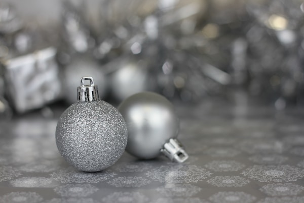 Two silver Christmas ornaments rest on a patterned surface, with one glittery and the other matte. The background is adorned with blurred silver decorations, creating a festive atmosphere.