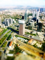 Aerial view of a modern cityscape featuring tall skyscrapers and a prominent, curved road network. The buildings are densely packed and vary in height and design, with the tallest structure in the foreground. Green spaces and infrastructure are visible, indicating a well-planned urban environment.