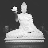 A serene white statue of a meditative figure, seated cross-legged and draped in flowing robes, holds a lotus flower in one hand. The intricate details of the robe's folds and the peaceful expression convey a sense of calm.