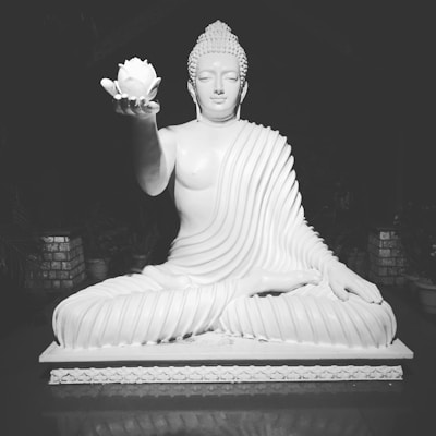 A serene white statue of a meditative figure, seated cross-legged and draped in flowing robes, holds a lotus flower in one hand. The intricate details of the robe's folds and the peaceful expression convey a sense of calm.