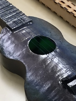 A close-up view of a dark-colored stringed instrument resembling a guitar, with a shiny surface and visible strings. The soundhole appears to have a green hue, and a patterned fretboard is partially seen in the background.