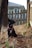A black dog with a white patch on its chest is sitting on a bed of dry leaves in a fenced backyard. It wears a red collar with a harness and ID tags, and its ears are perked up as it looks attentively to the side. A wooden fence and the edge of a house are visible in the background.