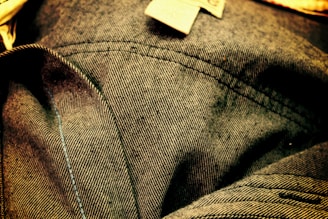 Close-up view of denim fabric showcasing detailed stitching and texture. The fabric appears wrinkled, with visible seam lines and a small fabric tag.