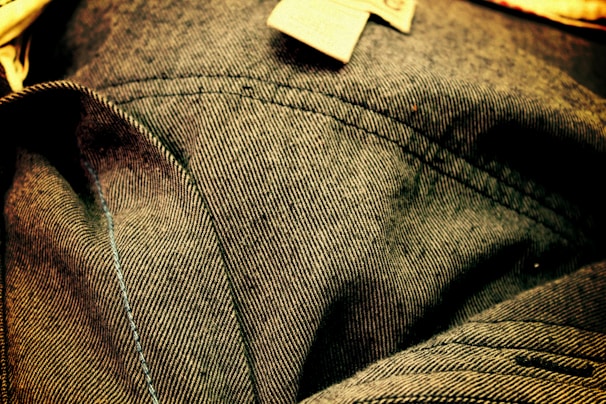 Close-up view of denim fabric showcasing detailed stitching and texture. The fabric appears wrinkled, with visible seam lines and a small fabric tag.