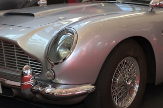 A classic car featuring a polished silver exterior with elegant curves and a vintage design. The focus is on the front part, highlighting the distinctive grille and rounded headlight, complemented by chrome detailing around the bumper and wheel hub.
