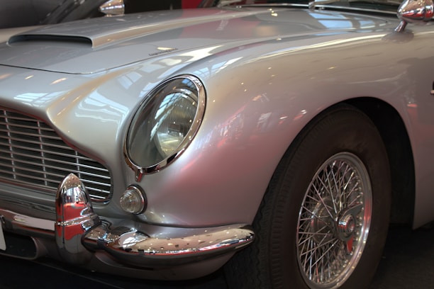 A classic car featuring a polished silver exterior with elegant curves and a vintage design. The focus is on the front part, highlighting the distinctive grille and rounded headlight, complemented by chrome detailing around the bumper and wheel hub.
