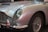 A classic car featuring a polished silver exterior with elegant curves and a vintage design. The focus is on the front part, highlighting the distinctive grille and rounded headlight, complemented by chrome detailing around the bumper and wheel hub.