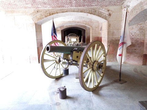 An old cannon with large spoked wheels is positioned inside a brick archway. The setting appears to be historic, with two flags - one of which resembles the United States flag - standing on either side of the cannon. The floor is made of stone, and the walls showcase a mix of stone and brickwork, adding to the historic ambiance.