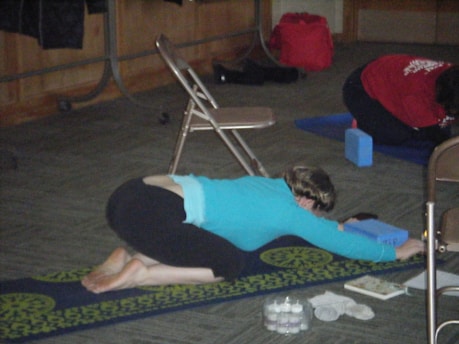 A person is practicing yoga on a mat in a child’s pose. The setting suggests an indoor space with chairs, yoga blocks, and other participants. In the background, a red bag and some personal items are visible.