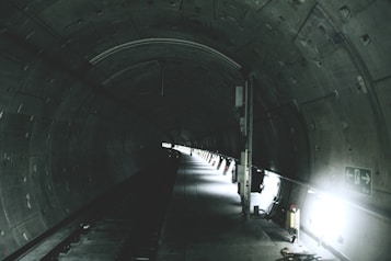 A dimly lit underground tunnel with a curved ceiling and industrial design. The walls are lined with panels and lights, casting a dim glow along the pathway. Emergency exit signs and utilities are visible along the sides.