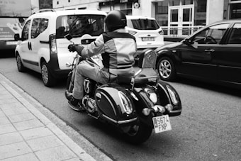 A person wearing a helmet and protective gear rides a motorcycle on a city street, surrounded by parked cars. The perspective is from behind, capturing the license plate and reflective chrome details of the motorcycle. The street is lined with vehicles, building facades, and a sidewalk.