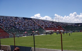 A large soccer stadium filled with spectators seated in the stands, wearing primarily blue clothing. The field is enclosed by a metal fence, and there are players on the grass pitch with a clear sky in the background.