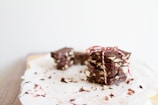 A stack of chocolate bark pieces tied with a red and white string, resting on a piece of parchment paper. The chocolate contains visible chunks of nuts and some crumbs are scattered around.