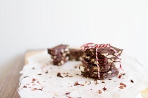 A stack of chocolate bark pieces tied with a red and white string, resting on a piece of parchment paper. The chocolate contains visible chunks of nuts and some crumbs are scattered around.