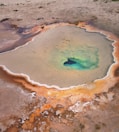 A natural hot spring with a vibrant color gradient ranging from deep turquoise at the center to rusty red and brown at the edges. The surrounding area is barren and rocky, highlighting the stark contrast of the colors in the geothermal pool.