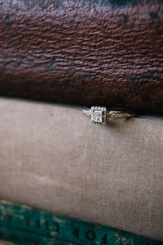A square-cut diamond ring is positioned on a textured, two-tone fabric surface with a leather book cover in the background, creating a contrast between luxury and natural materials.
