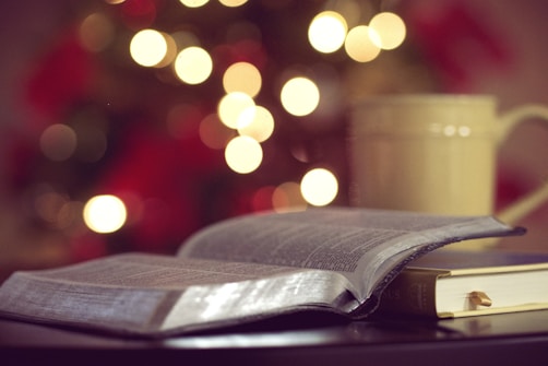 An open book lies on a table with the focus on the pages, creating a cozy and reflective atmosphere. In the background, there are blurred decorative lights and a large mug, enhancing the warmth of the scene.