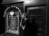 A woman in vintage attire and styled hair stands outside a venue with a decoration sign that reads 'Musical Diversion'. The marquee lists various jazz performances and dates. The scene combines elements of retro fashion with a music theme.