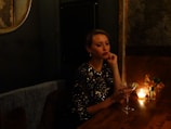 A woman in a sparkling black dress sits at a dimly lit wooden table, holding a martini glass with a lime garnish. A small candle and a vase with flowers add to the ambient mood. The setting has a luxurious and intimate feel, with textured wallpaper and a round mirror partially visible.