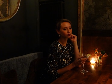 A woman in a sparkling black dress sits at a dimly lit wooden table, holding a martini glass with a lime garnish. A small candle and a vase with flowers add to the ambient mood. The setting has a luxurious and intimate feel, with textured wallpaper and a round mirror partially visible.