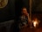 A woman in a sparkling black dress sits at a dimly lit wooden table, holding a martini glass with a lime garnish. A small candle and a vase with flowers add to the ambient mood. The setting has a luxurious and intimate feel, with textured wallpaper and a round mirror partially visible.