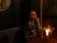 A woman in a sparkling black dress sits at a dimly lit wooden table, holding a martini glass with a lime garnish. A small candle and a vase with flowers add to the ambient mood. The setting has a luxurious and intimate feel, with textured wallpaper and a round mirror partially visible.