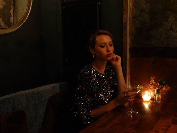 A woman in a sparkling black dress sits at a dimly lit wooden table, holding a martini glass with a lime garnish. A small candle and a vase with flowers add to the ambient mood. The setting has a luxurious and intimate feel, with textured wallpaper and a round mirror partially visible.