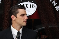 A man in a suit and tie is looking to the side, standing in front of a store sign that reads 'LEX 18' with text indicating it is a place for comfort food and diversion. The background includes a dark patterned curtain and a red lamp.