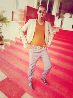 A person stands confidently on red carpeted steps in front of a building. They are wearing sunglasses, a blazer, an orange shirt, blue pants, and dress shoes.