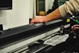 A person's hand operates a large black cutting machine, possibly a plotter or vinyl cutter. The machine is metallic and sleek, with a visible cutting mechanism and various electronic components. The setting seems to be an indoor workspace with blurred elements in the background.