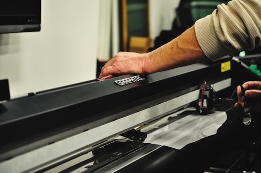Technician repairing a plotter machine with computer equipment in the background.