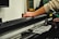 A person's hand operates a large black cutting machine, possibly a plotter or vinyl cutter. The machine is metallic and sleek, with a visible cutting mechanism and various electronic components. The setting seems to be an indoor workspace with blurred elements in the background.