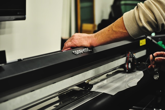 A person's hand operates a large black cutting machine, possibly a plotter or vinyl cutter. The machine is metallic and sleek, with a visible cutting mechanism and various electronic components. The setting seems to be an indoor workspace with blurred elements in the background.