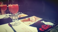 Two tall glasses filled with a red drink are placed on a table, accompanied by a menu from 'The Fat Chef'. The setting appears to be a restaurant with a dark and intimate ambiance. A napkin, a folded menu, and a glass of water are also visible on the table.