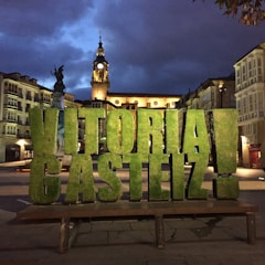 Large green letters spelling 'Vitoria-Gasteiz' are prominently displayed in an urban plaza. Behind the letters, an ornate clock tower is illuminated against a dark, cloudy evening sky. Surrounding the plaza are historical buildings with intricate facades and balconies.