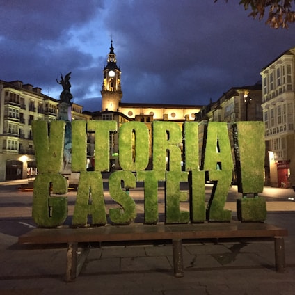 Large green letters spelling 'Vitoria-Gasteiz' are prominently displayed in an urban plaza. Behind the letters, an ornate clock tower is illuminated against a dark, cloudy evening sky. Surrounding the plaza are historical buildings with intricate facades and balconies.