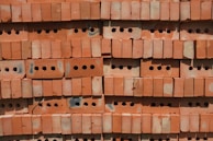A stack of red perforated bricks arranged neatly in horizontal rows. Each brick has multiple round holes, providing a uniform, textured pattern. The surface shows signs of slight discoloration and wear, possibly from exposure to weather.