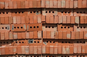 A stack of red perforated bricks arranged neatly in horizontal rows. Each brick has multiple round holes, providing a uniform, textured pattern. The surface shows signs of slight discoloration and wear, possibly from exposure to weather.