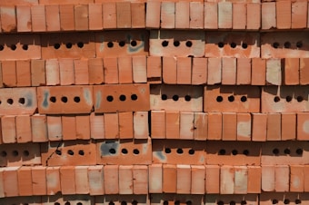 A stack of red perforated bricks arranged neatly in horizontal rows. Each brick has multiple round holes, providing a uniform, textured pattern. The surface shows signs of slight discoloration and wear, possibly from exposure to weather.