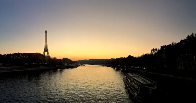 A serene view of the Eiffel Tower at sunset with the Seine River flowing gently beneath a clear, gradient sky transitioning from orange to blue. Trees line the riverbank on both sides, silhouetted against the light. Boats are docked along the river, and the overall scene is calm and picturesque.