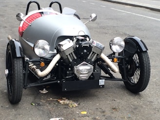 A vintage-style three-wheeled vehicle with a prominent front engine and open cockpit. The vehicle features large round headlights, a black wheel covering, and a retro aesthetic. The body is sleek with a silver color and red quilted upholstery visible inside. It is parked on a slightly worn urban street.