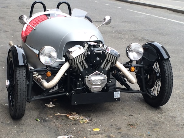 A vintage-style three-wheeled vehicle with a prominent front engine and open cockpit. The vehicle features large round headlights, a black wheel covering, and a retro aesthetic. The body is sleek with a silver color and red quilted upholstery visible inside. It is parked on a slightly worn urban street.