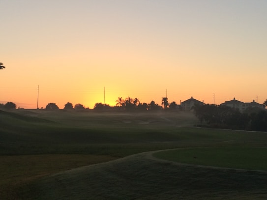The image captures a serene sunset over a landscape featuring a well-maintained golf course and silhouetted buildings and palm trees in the distance. The sky transitions from a deep blue near the horizon to a warm, glowing orange as the sun sets. Mist gently hovers over the grassy areas.