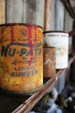 An old, rusty can of liquid rubber with vivid red paint dripping from the top stands prominently on a wooden shelf. There are other similarly aged cans visible in the background, out of focus. The setting appears to be a dusty and ambient space, possibly an old workshop or garage, with a rustic and vintage atmosphere.